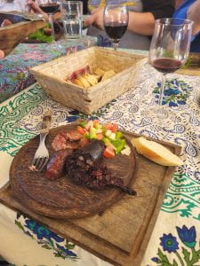 A photo of a round wooden plate holding a broken link of morcilla, a piece of chorizo, a piece of white baguette, and an ensalada criolla. There is a glass of half-drunk red wine, and there are belongings of other people gathered at the table in the background.