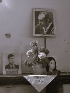 A B/W photo of a memorial altar, with flowers and three photos of "disappeared" photos. A draped cloth says "nunca mas" below the flowers.